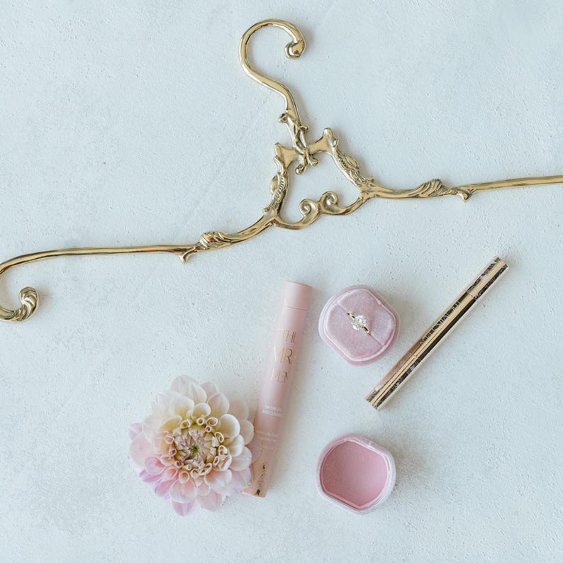 K&ouml;nnte beinhalten: Ein goldener, verzierter Kleiderb&uuml;gel mit einer rosafarbenen Ringbox, einer rosafarbenen Ringbox mit einem Diamantring, einer rosafarbenen Lippenbalsamtube, einer rosafarbenen Blume und einem goldenen Eyeliner-Stift.