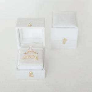 May include: Two gold rings in a white ring box. The top ring has a large oval-shaped clear stone. The bottom ring has a delicate design with small clear stones.