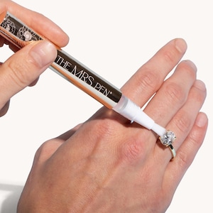 May include: A gold and white pen labeled "THE MRS. PEN" is being used to clean a diamond ring on a person's finger.