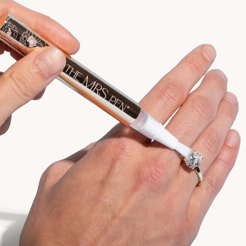 May include: A gold and white pen labeled "THE MRS. PEN" is being used to clean a diamond ring on a person's finger.