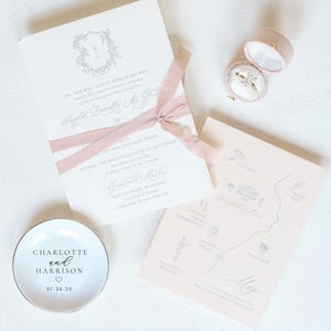 May include: Wedding invitation suite featuring a cream-colored invitation with a floral crest and pink ribbon. Includes a map, ring box, and a small white dish with the names "Charlotte and Harrison" and the date 07-28-26.