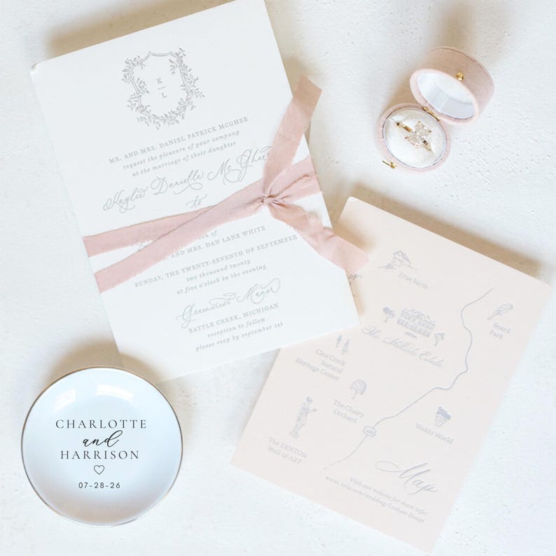 May include: Wedding invitation suite featuring a cream-colored invitation with a floral crest and pink ribbon. Includes a map, ring box, and a small white dish with the names "Charlotte and Harrison" and the date 07-28-26.