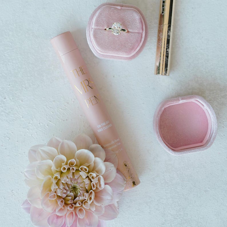 K&ouml;nnte beinhalten: Eine rosafarbene Samt-Ringbox mit einem Diamantring darin, eine rosafarbene Tube Handcreme mit dem Text "The Mrs. Pen" darauf, eine rosafarbene Samtbox und eine rosafarbene Blume.