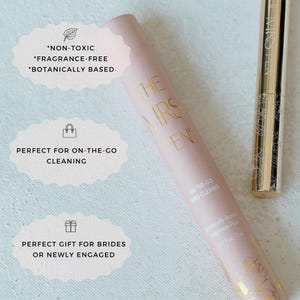 K&ouml;nnte beinhalten: Eine rosa und goldene Tube mit dem Text "The Mrs. Pen" und "On-the-Go Ring Cleaner". Die Tube ist als ungiftig, parf&uuml;mfrei und auf pflanzlicher Basis gekennzeichnet. Die Tube befindet sich neben einem goldenen Stift mit dem Text "The Mrs. Pen".