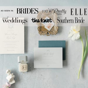 Op de afbeelding: Bruiloft briefpapier set met een uitnodiging, antwoordkaart en envelop in zachte witte en blauwe tinten. Een kleine ringdoos en een decoratief dienblad met postzegels zijn ook inbegrepen. Een witte bloem en een kleine buste.