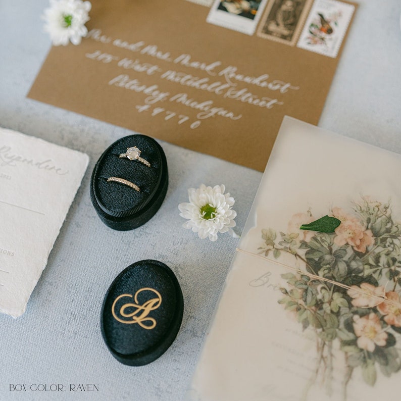 May include: Two black velvet ring boxes with gold lettering. One box has a single ring with a large diamond and a smaller band. The other box has the letter "A" engraved on the top. The boxes are on a light blue surface with a white flower and a brown envelope.