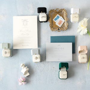 May include: A sophisticated wedding stationery set with a main invitation, a reply card, and a teal envelope. Several ring boxes in various colours, including black, green, and pink, are open, displaying diamond rings. A gold tray with stamps and a white flower complete the arrangement.