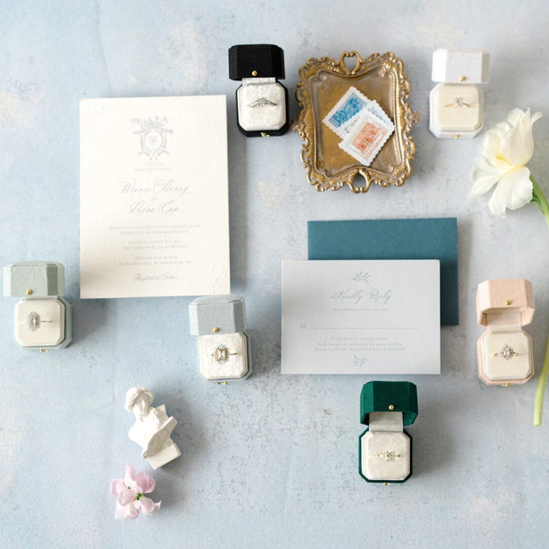 May include: A sophisticated wedding stationery set with a main invitation, a reply card, and a teal envelope. Several ring boxes in various colours, including black, green, and pink, are open, displaying diamond rings. A gold tray with stamps and a white flower complete the arrangement.