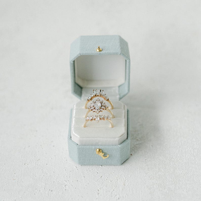 Op de afbeelding: Een open, lichtblauwe achthoekige ringendoos met een gouden sluiting, met drie gouden ringen met sprankelende edelstenen. De ringen liggen op een wit fluwelen interieur, wat een luxe uitstraling cre&euml;ert.