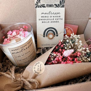 May include: A gift box containing a soy wax candle in a glass jar with pink dried flowers, a gold-toned sun charm bracelet on a card with the text 'maitresse MERCI D'AVOIR ENSOLEILL&Eacute; CETTE BELLE ANN&Eacute;E', and a bouquet of pink and white dried flowers.