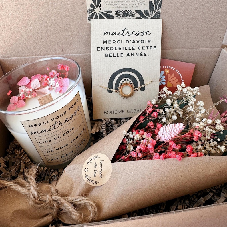May include: A gift box containing a soy wax candle in a glass jar with pink dried flowers, a gold-toned sun charm bracelet on a card with the text 'maitresse MERCI D'AVOIR ENSOLEILL&Eacute; CETTE BELLE ANN&Eacute;E', and a bouquet of pink and white dried flowers.