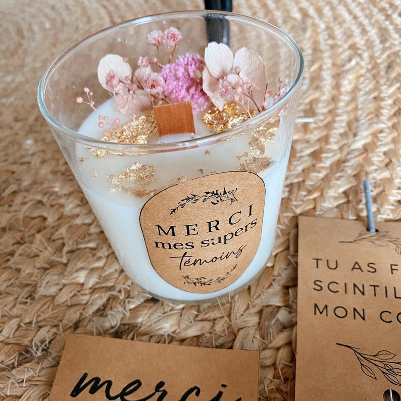 May include: A white candle with a brown label that says "Merci mes supers temoins". The candle is decorated with dried flowers and gold flakes.