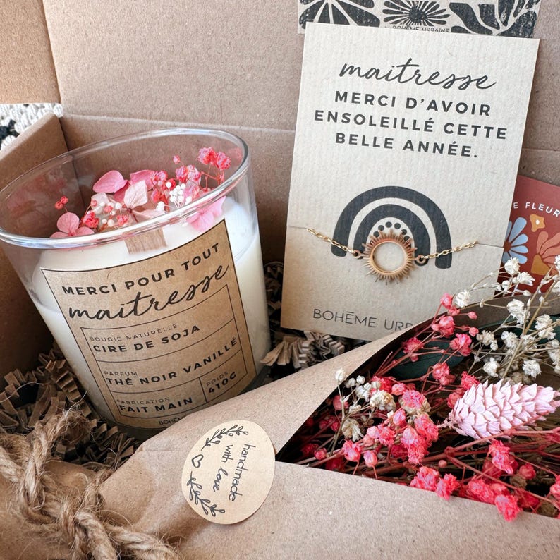 May include: A gift box containing a soy wax candle in a glass jar with pink dried flowers, labeled Merci pour tout maitresse, and a gold-tone sun charm bracelet on a card that reads maitresse MERCI D'AVOIR ENSOLEILL&Eacute; CETTE BELLE ANN&Eacute;E.