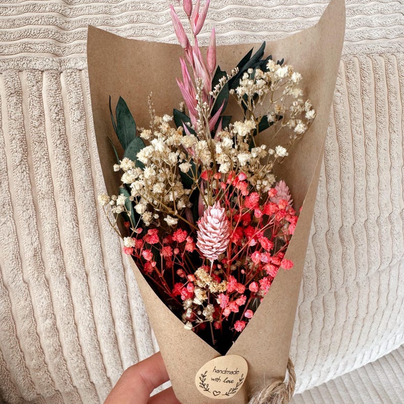 May include: A small bouquet of dried flowers wrapped in brown kraft paper. The arrangement includes white baby's breath, pink dyed baby's breath, dark green leaves, and tall pink stalks. A round sticker on the paper reads handmade with love.