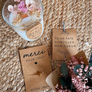 May include: A brown card with the text "merci pour tout" and a small heart. The card is decorated with a gold star and the text "BOHEME URBAIN". A second brown card with the text "TU AS FAIS SCINTILLER MON COEUR" is visible in the background. A clear glass candle jar with dried flowers and gold flakes is also visible in the background.