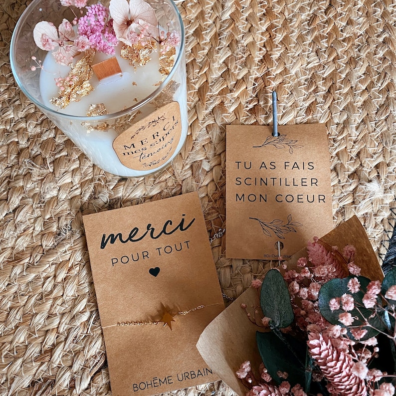 May include: A brown card with the text "merci pour tout" and a small heart. The card is decorated with a gold star and the text "BOHEME URBAIN". A second brown card with the text "TU AS FAIS SCINTILLER MON COEUR" is visible in the background. A clear glass candle jar with dried flowers and gold flakes is also visible in the background.