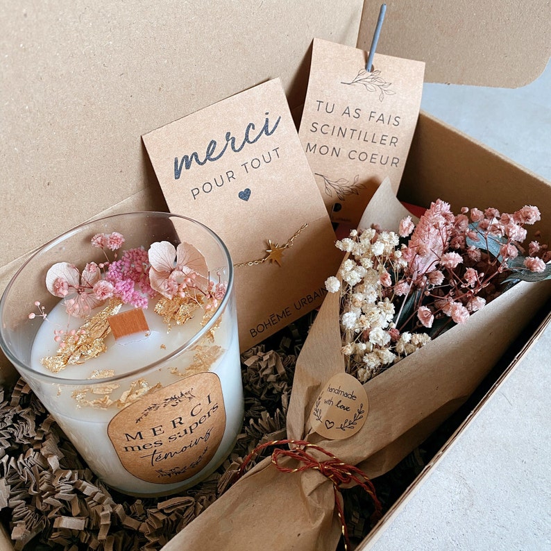 May include: A gift box filled with a white candle with dried flowers and gold flakes, a small bouquet of dried flowers wrapped in brown paper, a gold necklace with a star charm, and two cards. One card says "merci pour tout" and the other says "tu as fais scintiller mon coeur".