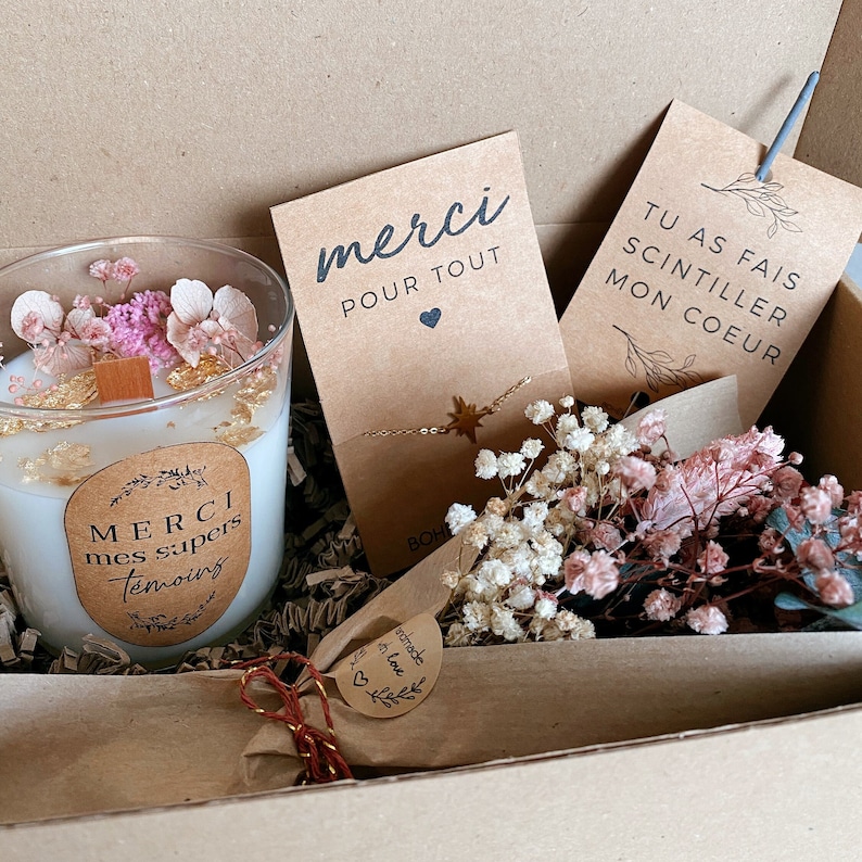 May include: A gift box filled with a variety of items, including a candle with dried flowers, a necklace, a small bouquet of dried flowers, and several cards with handwritten messages. The cards say "merci pour tout" and "tu as fais scintiller mon coeur".