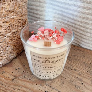 May include: A clear glass candle with white soy wax and a wooden wick, topped with dried pink and red flowers. The label features the text "MERCI POUR TOUT maitresse", "CIRE DE SOJA", "THE NOIR VANILLE", and "FAIT MAIN 410G" in black ink on a tan background.