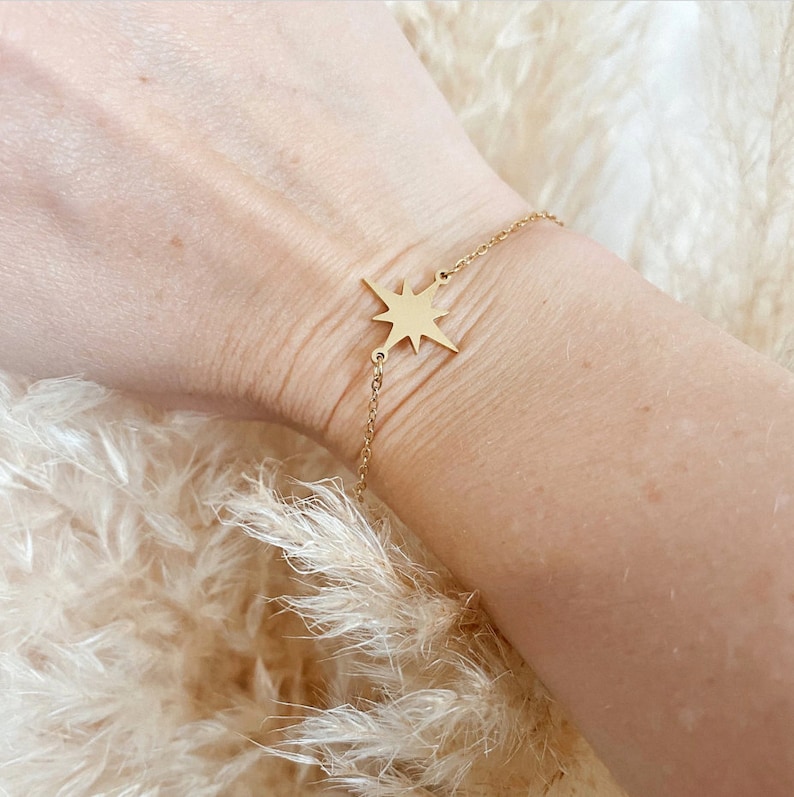 May include: A gold chain bracelet with a starburst charm. The bracelet is on a person's wrist.