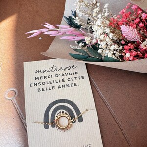 May include: A gold-toned sunburst charm bracelet attached to a rectangular card featuring a black rainbow graphic and the French text: maitresse MERCI D'AVOIR ENSOLEILL&Eacute; CETTE BELLE ANN&Eacute;E. The card is positioned below a bouquet of dried flowers.