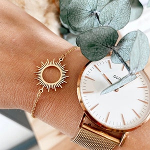 May include: A gold-toned chain bracelet featuring a central sunburst charm with radiating rays. The bracelet is worn alongside a gold-toned wristwatch with a white face, thin gold markers, and a fine mesh metal band, accented by green leaves.