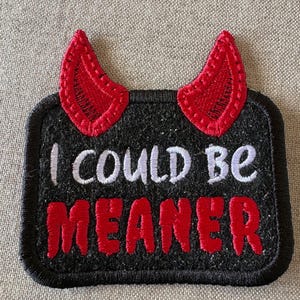 May include: A rectangular black embroidered patch featuring two red devil horns at the top. The text I COULD BE is stitched in white thread, while the word MEANER is stitched in bold red thread. The entire patch has a thick black stitched border.
