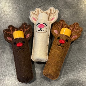 May include: Three felt reindeer lip balm holders. One is white with a pink nose and brown eyes. The other two are brown with red noses and brown eyes.