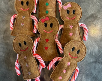 May include: Six gingerbread people ornaments with candy cane arms. The gingerbread people are brown with red and white candy cane arms. The gingerbread people have embroidered faces and details.