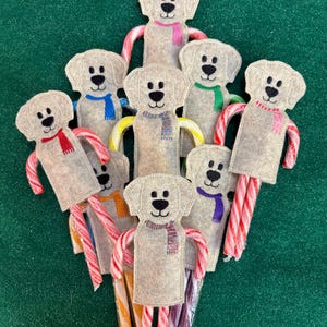 May include: A collection of felt dog ornaments with candy cane arms and colorful scarves. The dogs have black button eyes and are arranged on a green background. Each dog is unique with different scarf colors.