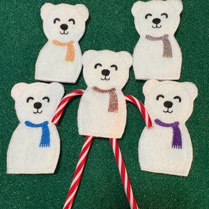May include: Five white felt polar bear ornaments with black stitched eyes and smiles, each wearing a colorful scarf. The bears are arranged around two red and white striped candy canes, creating a festive holiday decoration.