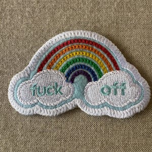 May include: An embroidered patch featuring a colorful rainbow arching over two white clouds. The left cloud has the word "fuck" stitched in light blue thread, and the right cloud has the word "off" stitched in the same light blue thread.