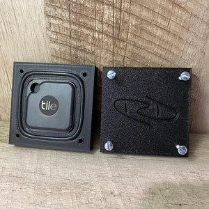 May include: A black square case with a Tile tracker inside, next to a matching black cover. The cover has the letters "TZL" engraved on it and is secured with four silver screws. The Tile tracker is black with the word "tile" in white.
