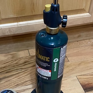 May include: A green propane torch with a black nozzle and a yellow control knob. The torch is labeled "ZOMATIC" and "PROPANE". Soldering paste and solder are visible in the foreground.