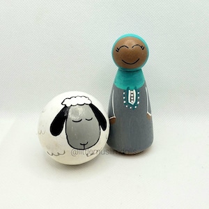 May include: A hand-painted wooden doll wearing a teal headscarf and gray dress, standing next to a white wooden sheep with a gray face.