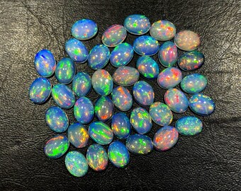 Puede incluir: Una colección de ópalos de forma ovalada con colores iridiscentes, incluyendo azul, verde y rojo. Los ópalos están dispuestos en un montón suelto sobre una superficie de cuero marrón oscuro.