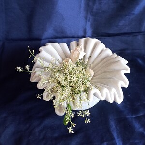 May include: White marble bowl with a ruffled edge, filled with white flowers and seashells. The bowl is on a dark blue background.