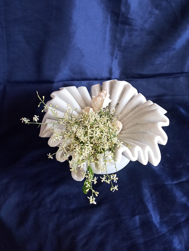 May include: White marble bowl with a ruffled edge, filled with white flowers and seashells. The bowl is on a dark blue background.