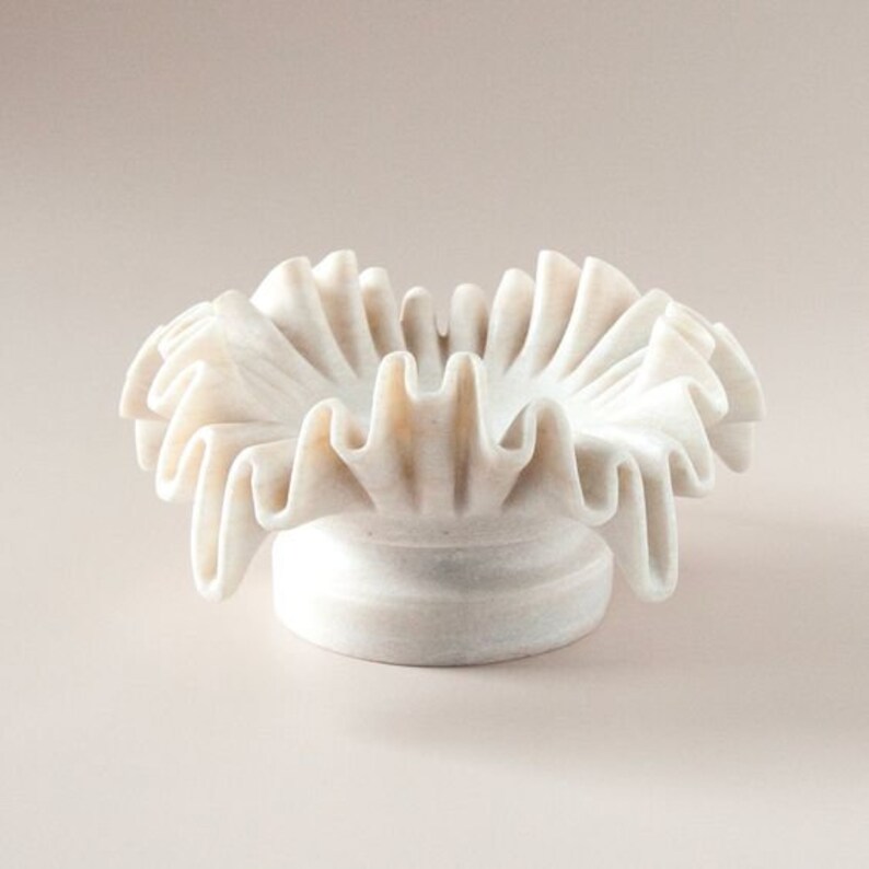 May include: A white marble bowl with a ruffled edge. The bowl is shaped like a flower and has a smooth, polished finish.
