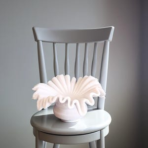 May include: A white ceramic vase with a ruffled, organic shape sits on a gray wooden chair. The vase is a modern, sculptural design.