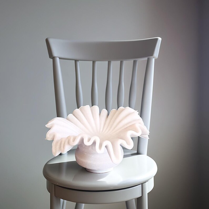May include: A white ceramic vase with a ruffled, organic shape sits on a gray wooden chair. The vase is a modern, sculptural design.