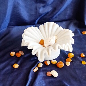 May include: A white ceramic bowl shaped like a flower with ruffled edges. The bowl is filled with small orange and brown stones and a white seashell. The bowl is sitting on a blue fabric surface.