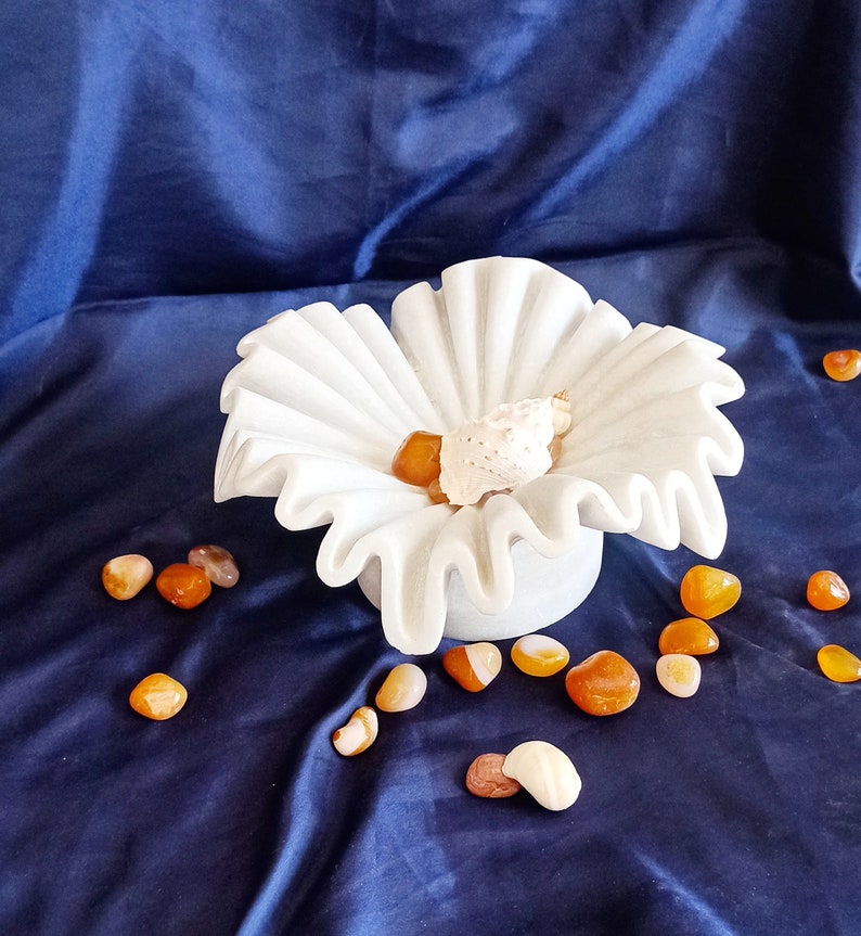 May include: A white ceramic bowl shaped like a flower with ruffled edges. The bowl is filled with small orange and brown stones and a white seashell. The bowl is sitting on a blue fabric surface.