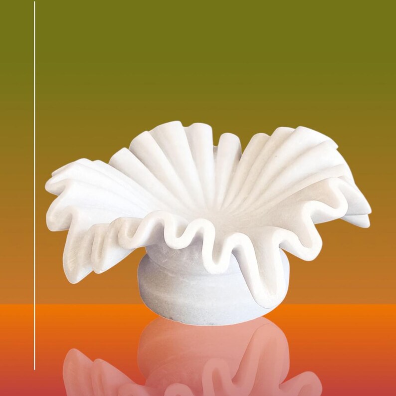 May include: A white marble decorative bowl with a ruffled, flower-like design. The bowl is sitting on a reflective orange surface.
