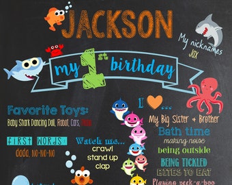 May include: A chalkboard style birthday poster for a boy named Jackson turning one year old. The poster features a blue and white baby shark character, a red octopus, and a red crab. The poster includes details about Jackson's first year, including his favourite toys, first words, height, weight, and favourite foods. The poster also includes the text "My nicknames: JOX".