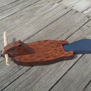 May include: A wooden beaver figurine with a brown body, dark brown head, and a blue tail. The beaver holds a light-colored wooden stick in its mouth. The figurine is painted and sits on a wooden surface.