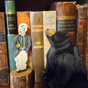 May include: A wooden carving of a black bear sits next to a wooden figure of a man in a suit, standing on a tree stump. Books with titles like "William Faulkner" and "Light in August" are in the background.