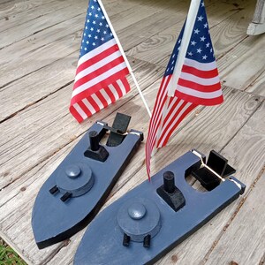 May include: Two wooden toy boats painted blue with black accents, each featuring a central turret and a small black structure. Two American flags are placed behind the boats, with red and white stripes and a field of white stars.