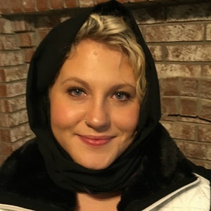 May include: A person with blonde hair and blue eyes wearing a black head covering and a black fur-trimmed jacket. The person is smiling in front of a brick wall.