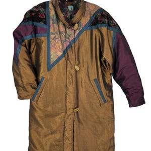 Op de afbeelding: Een vintage lange jas in tinten brons, paars en bloempatronen. De jas heeft een hoge kraag, houten knopen en een uniek colorblock-ontwerp met blauwe biezen. De mouwen zijn effen paars en de schouders hebben bloemaccenten.